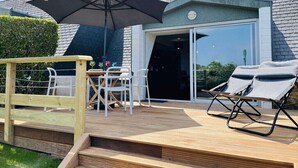 Een houten terras met buitenmeubilair, een grote parasol en een glazen deur die naar een gebouw leidt.
