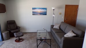 A living room with a sofa, armchair, glass coffee table, and a painting of a sunset. - Quaint updated apartment overlooking the Bay (Washburn)