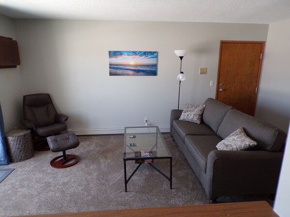 A living room with a sofa, armchair, glass coffee table, and a painting of a sunset.