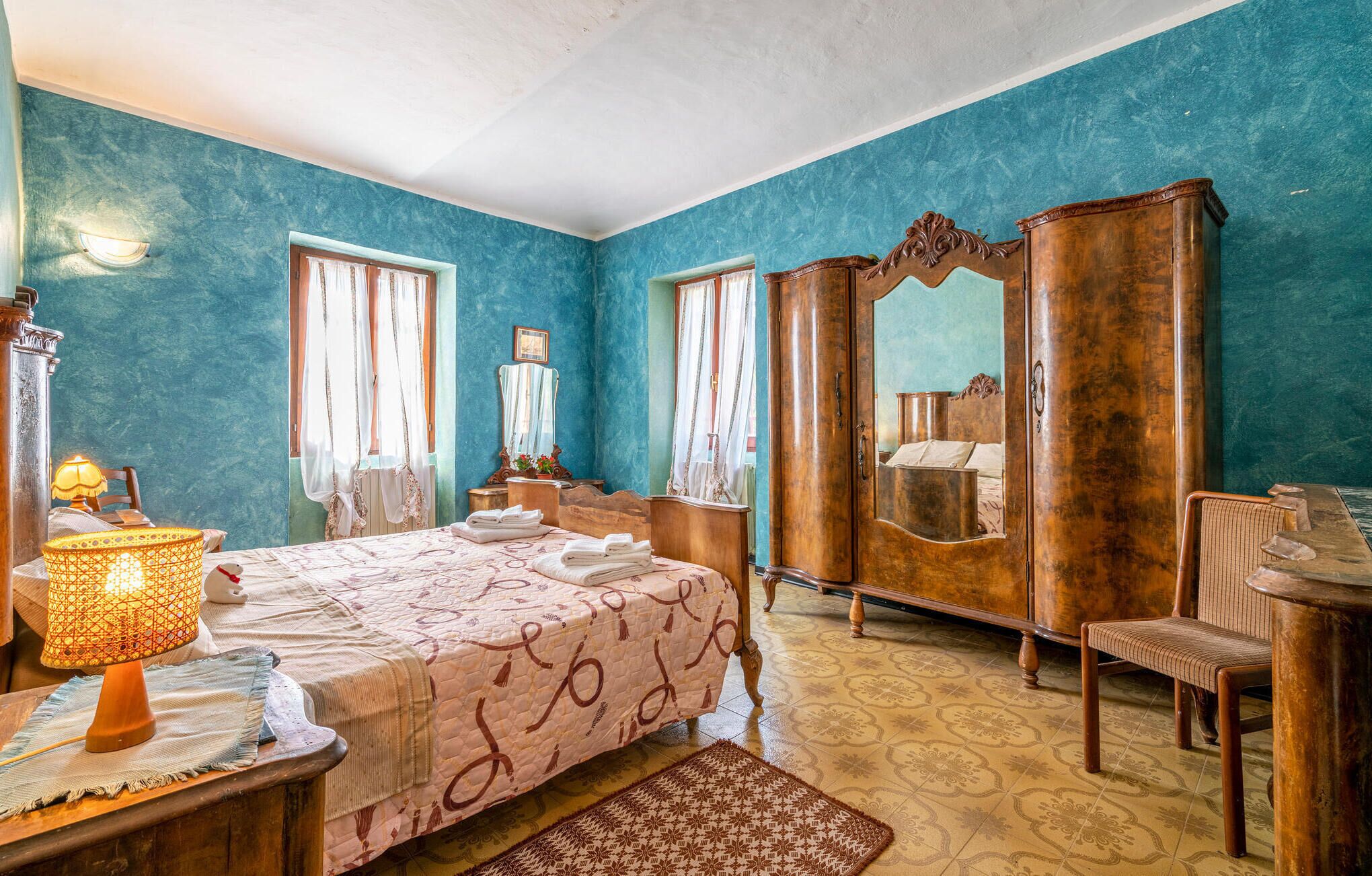 A bedroom with a bed, a wooden wardrobe, a mirror, a chair, and a lamp.