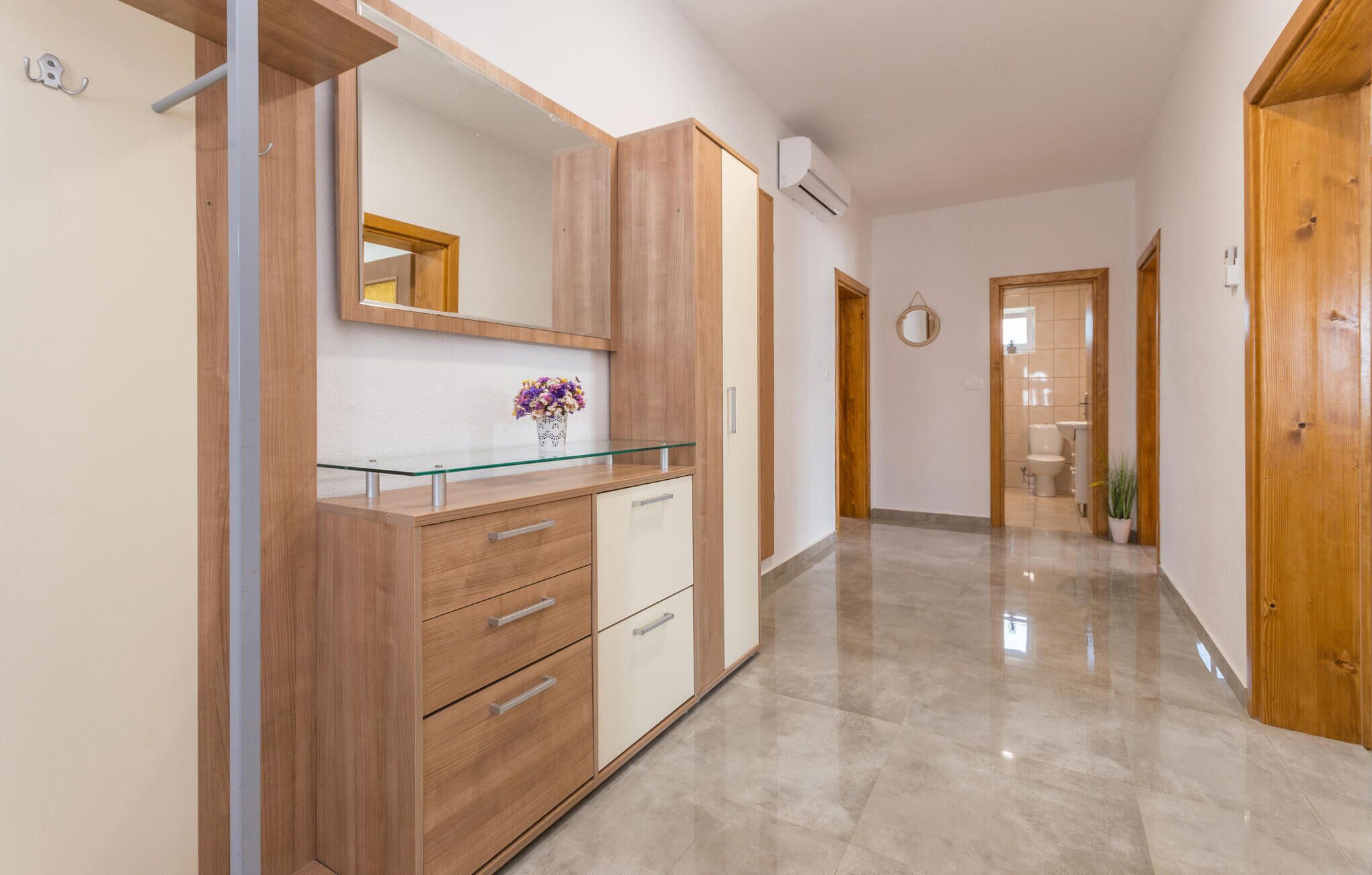 A hallway with a wooden cabinet, a glass-top desk, and a mirror. There is a potted plant and a door leading to a bathroom.
