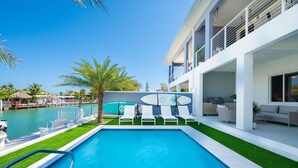A modern pool area with a clear blue pool, white lounge chairs, and a white building with balconies. - Canal Front with Cabana Club Privileges! Paddleboards, Bikes, Kayak in KCB! (Key Colony Beach)