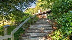 Una escalera de madera rodeada de exuberante vegetación, que conduce a una zona cubierta con un muro de ladrillo y una chimenea.