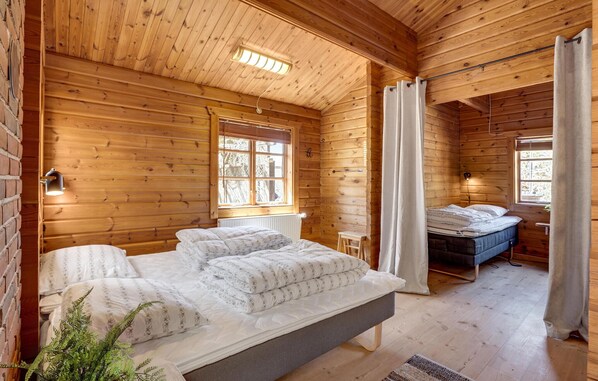 Een slaapkamer in een blokhut met een bed, een klein krukje en een raam met gordijnen.