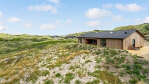 Eine Holzhütte mit einer großen Terrasse, umgeben von grasbewachsenen Dünen und Hügeln.