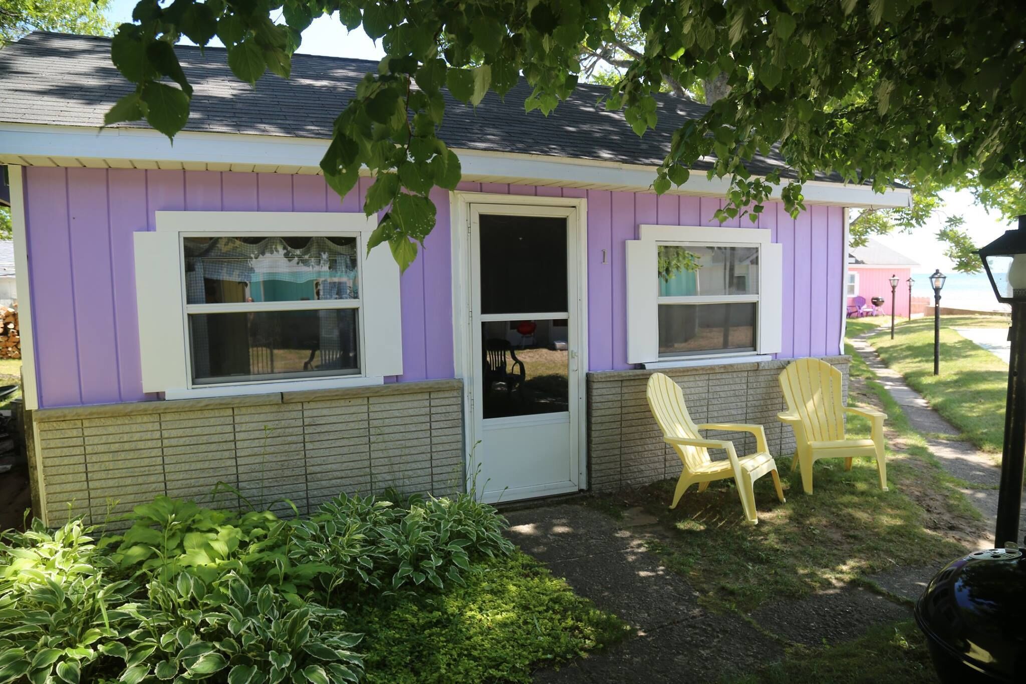 A small, single-story building with a purple exterior and white trim. It features two windows with white frames, a front door, and a small porch area with two yellow Adirondack chairs.