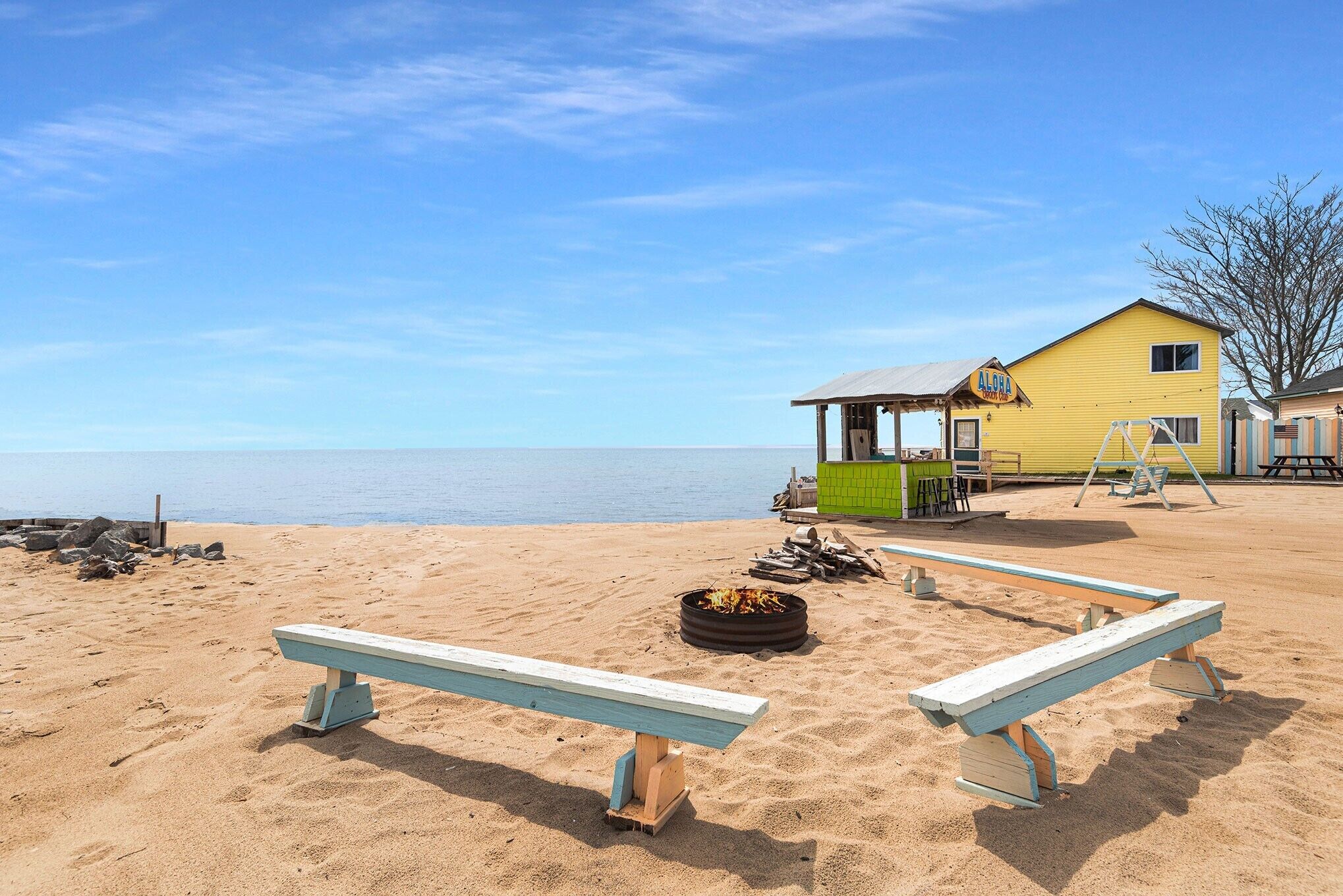 A beach with a yellow building, a fire pit, and wooden benches.