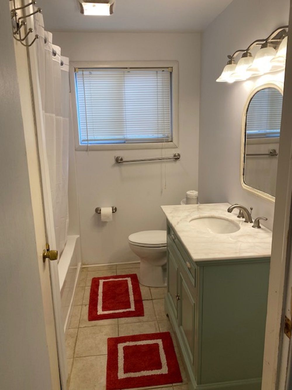 A small bathroom with a toilet, sink, and shower. There are two red rugs on the floor.
