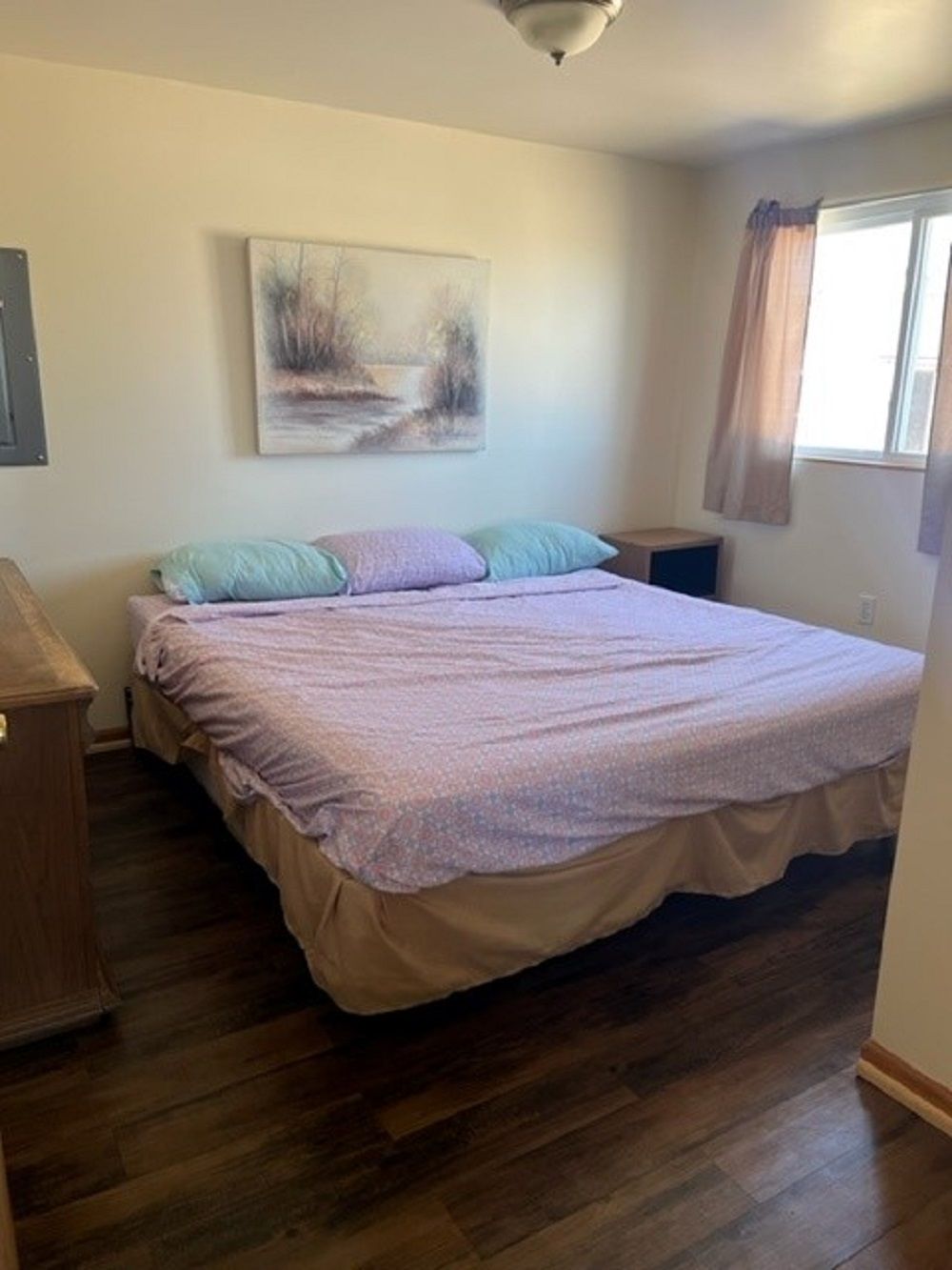 A bedroom with a bed, a nightstand, a lamp, a painting on the wall, and a window with curtains.
