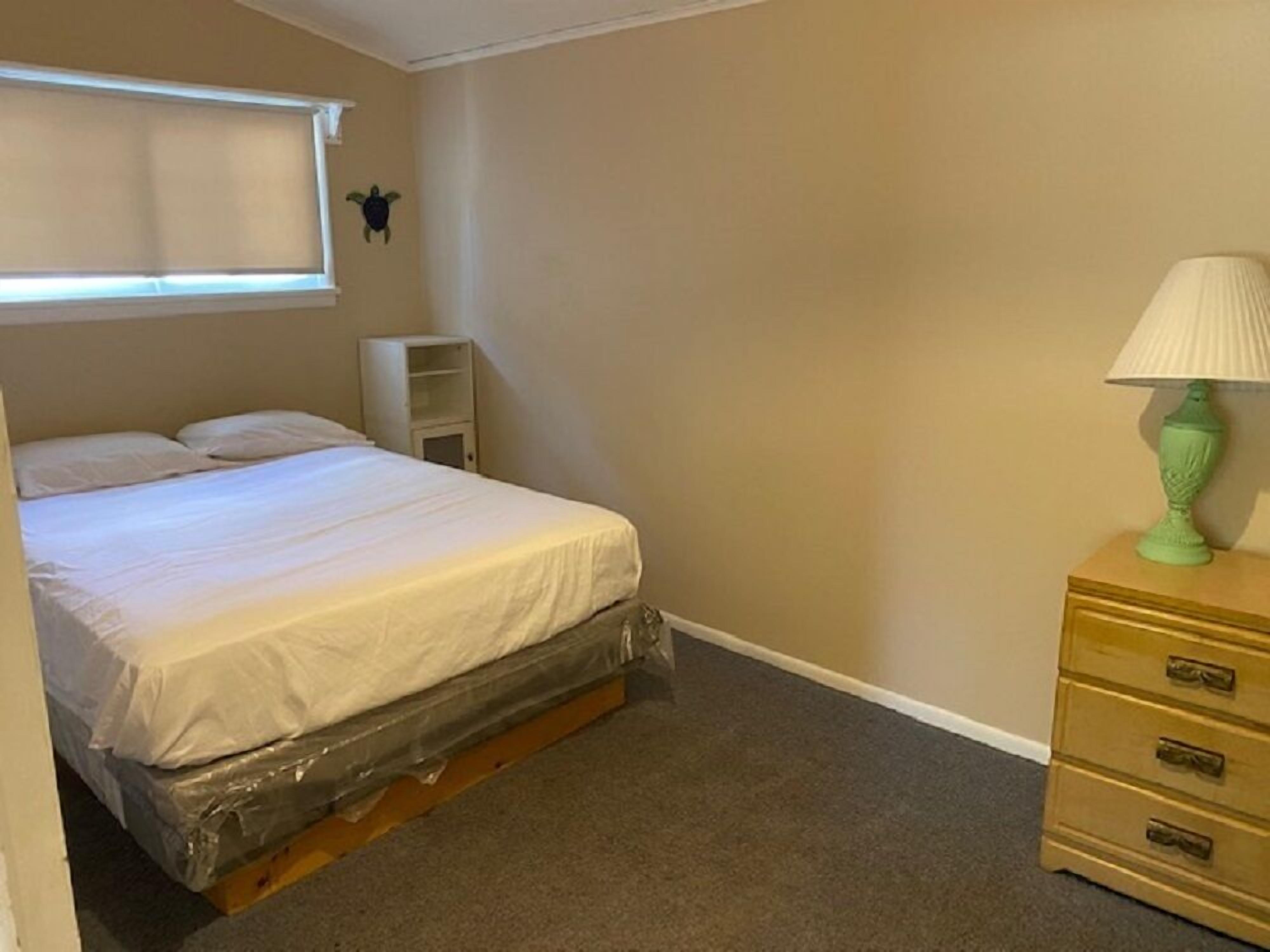A bedroom with a bed, a nightstand, a lamp, a small shelf, and a window with blinds.