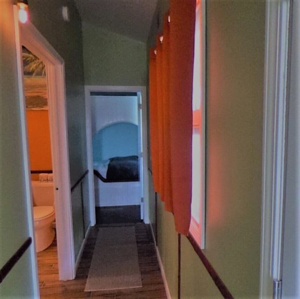 Un couloir étroit avec un plancher en bois, une porte blanche au fond et une fenêtre ornée de rideaux orange.