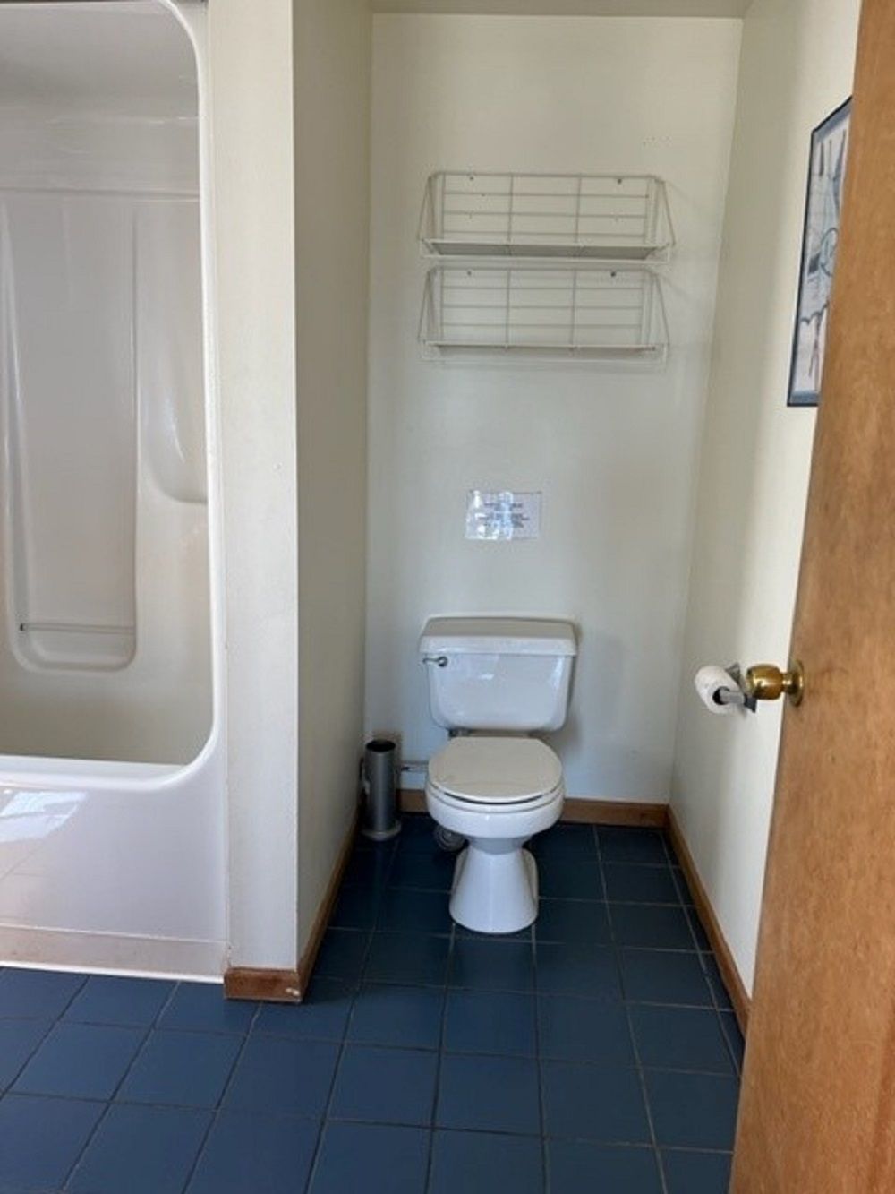 Un baño pequeño con inodoro, una bañera blanca y cortina de ducha. Hay dos repisas sobre el inodoro.