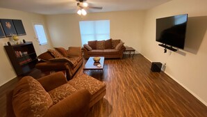 Uma sala de estar com piso de madeira, dois sofás, uma mesa de centro, uma estante de livros, uma TV de tela plana e um ventilador de teto.