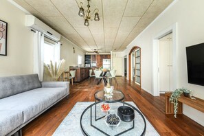 Un salon moderne avec un canapé, une table basse en verre sur laquelle on voit des fruits, un téléviseur et, au fond, une cuisine.