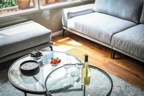 Une table à plateau en verre sur laquelle se trouvent un saladier de raisins, un saladier de fraises, des verres à vin et une bouteille de vin, dans un salon avec un canapé gris et une fenêtre.