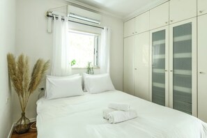 Une chambre à coucher avec un lit, des rideaux blancs, une fenêtre, un vase contenant des plumes d’herbe des pampas et une armoire.