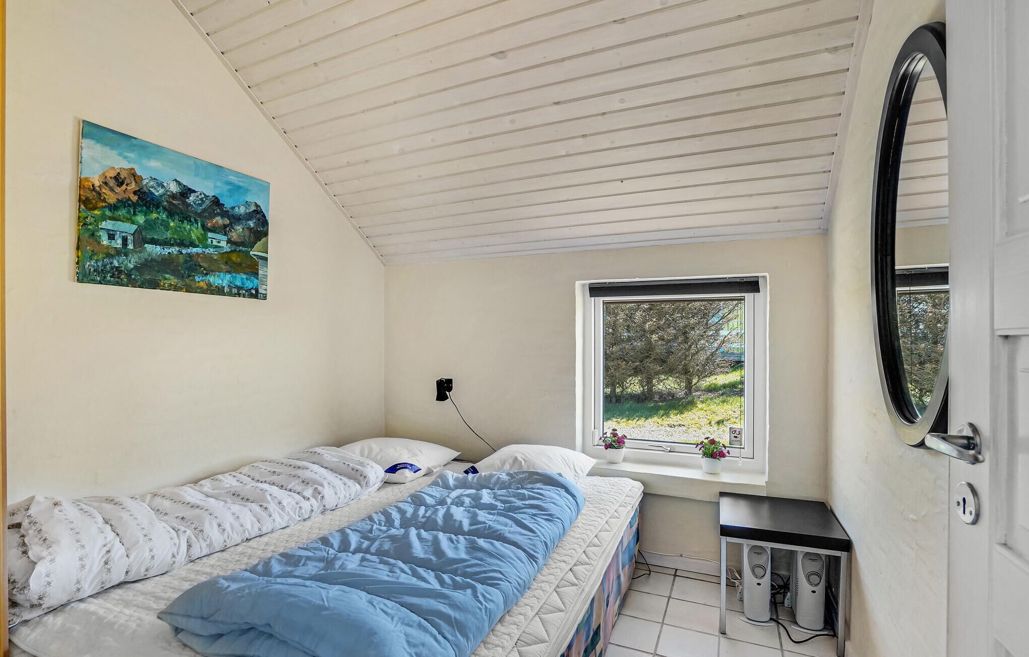 Ein Schlafzimmer mit Bett, einem Fenster mit Blick ins Grüne, einem Bild an der Wand und einer Tür mit Spiegel.