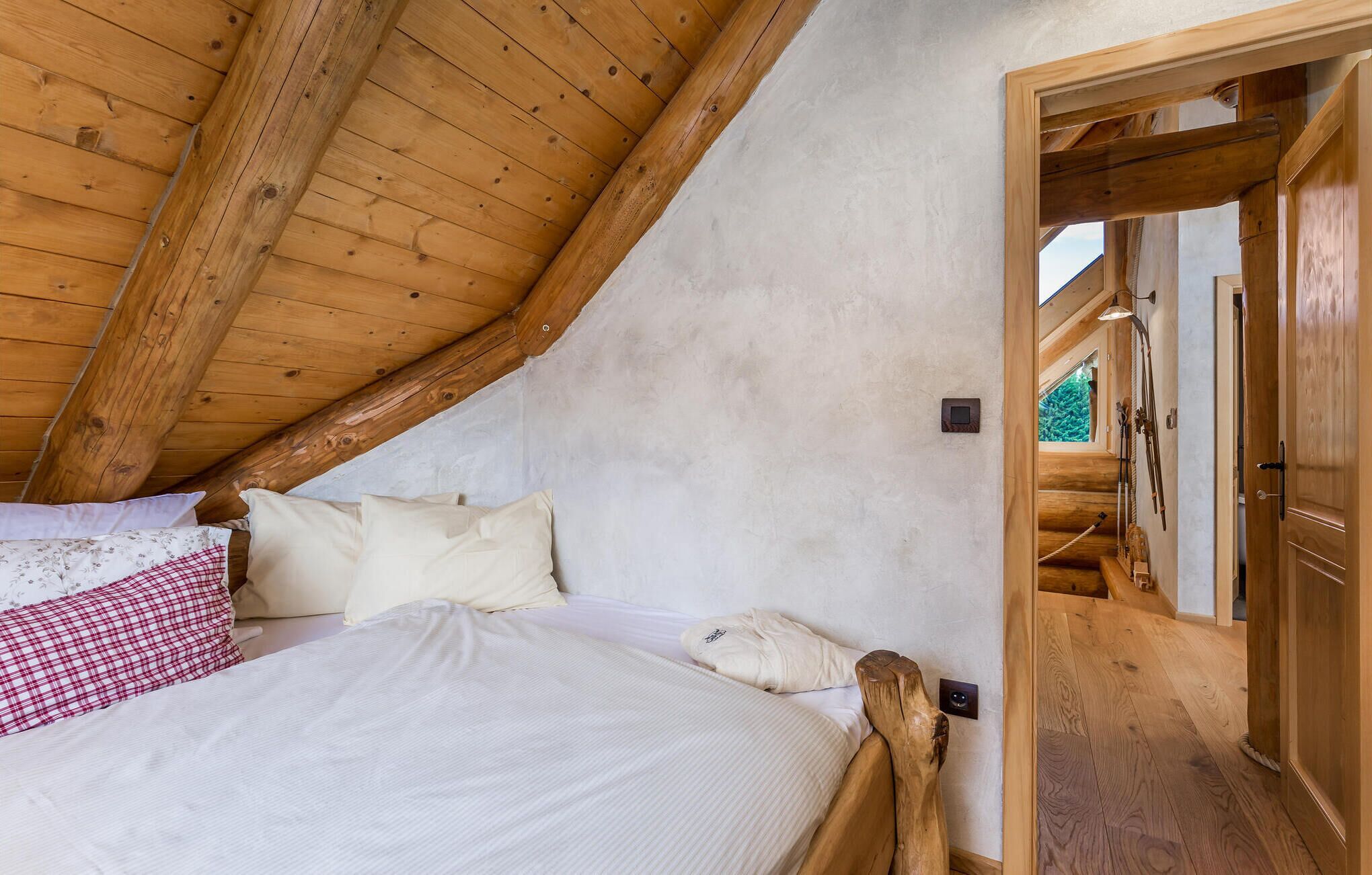 A bedroom with a sloped ceiling, wooden beams, a bed with white bedding, and a view of a wooden staircase leading to an outdoor area.