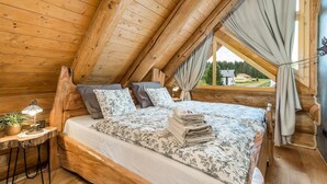 A cozy attic bedroom with a wooden sloped ceiling, a bed with floral bedding, a small table with a lamp, and a view of the outside. - Amazing home in Hlevci with sauna (Hlevci)