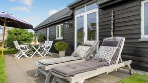 A wooden house with a patio area featuring two lounge chairs with cushions, a table with chairs, and a potted plant.