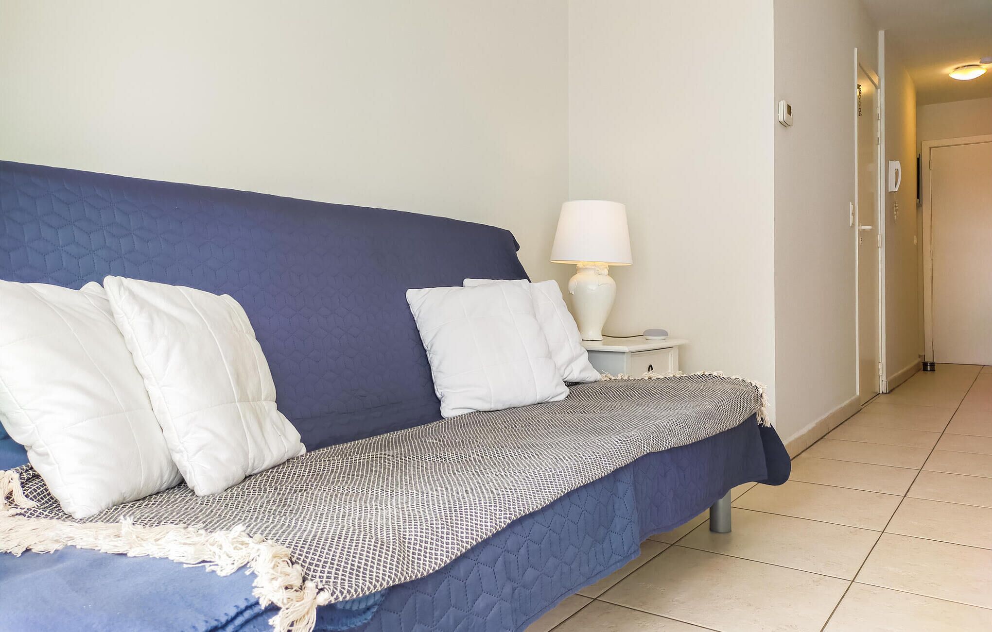 Une chambre avec un lit bleu, des oreillers blancs, une couverture à motifs, une table de chevet avec une lampe et une porte visible au fond.