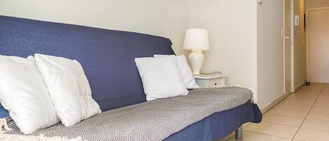 Une chambre avec un lit bleu, des oreillers blancs, une couverture à motifs, une table de chevet avec une lampe et une porte visible au fond.