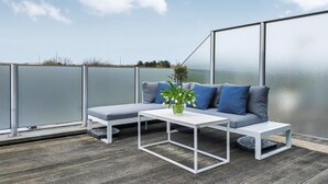 Un balcón con un sofá gris, una mesa de centro blanca y un jarrón con flores.
