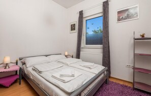 A bedroom with a bed, bedside lamps, a window with curtains, a radiator, and a shelf with a decorative item.