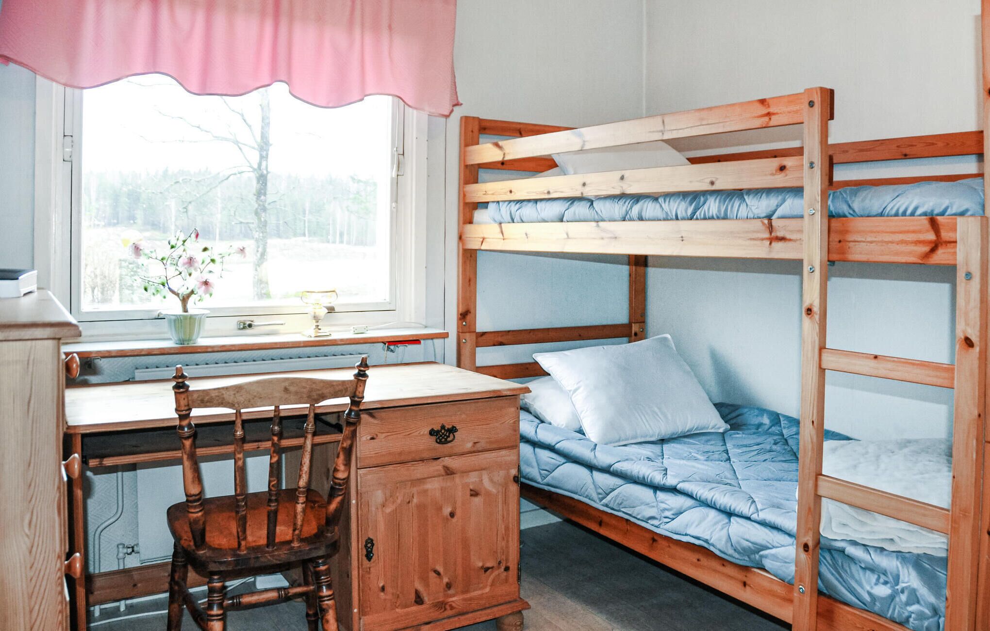 A bunk bed with a desk and chair, a window with a view of trees, and a pink curtain.