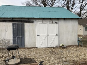Il s’agit d’un petit bâtiment d’un seul étage, doté d’un toit en métal ondulé et d’une façade blanche. On y trouve une grande porte de garage blanche et un petit espace porche avec un barbecue et un pot de fleurs.