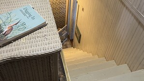 Un escalier, une chaise en osier sur laquelle repose un livre, une plaque décorative au mur et un papier peint à motifs.