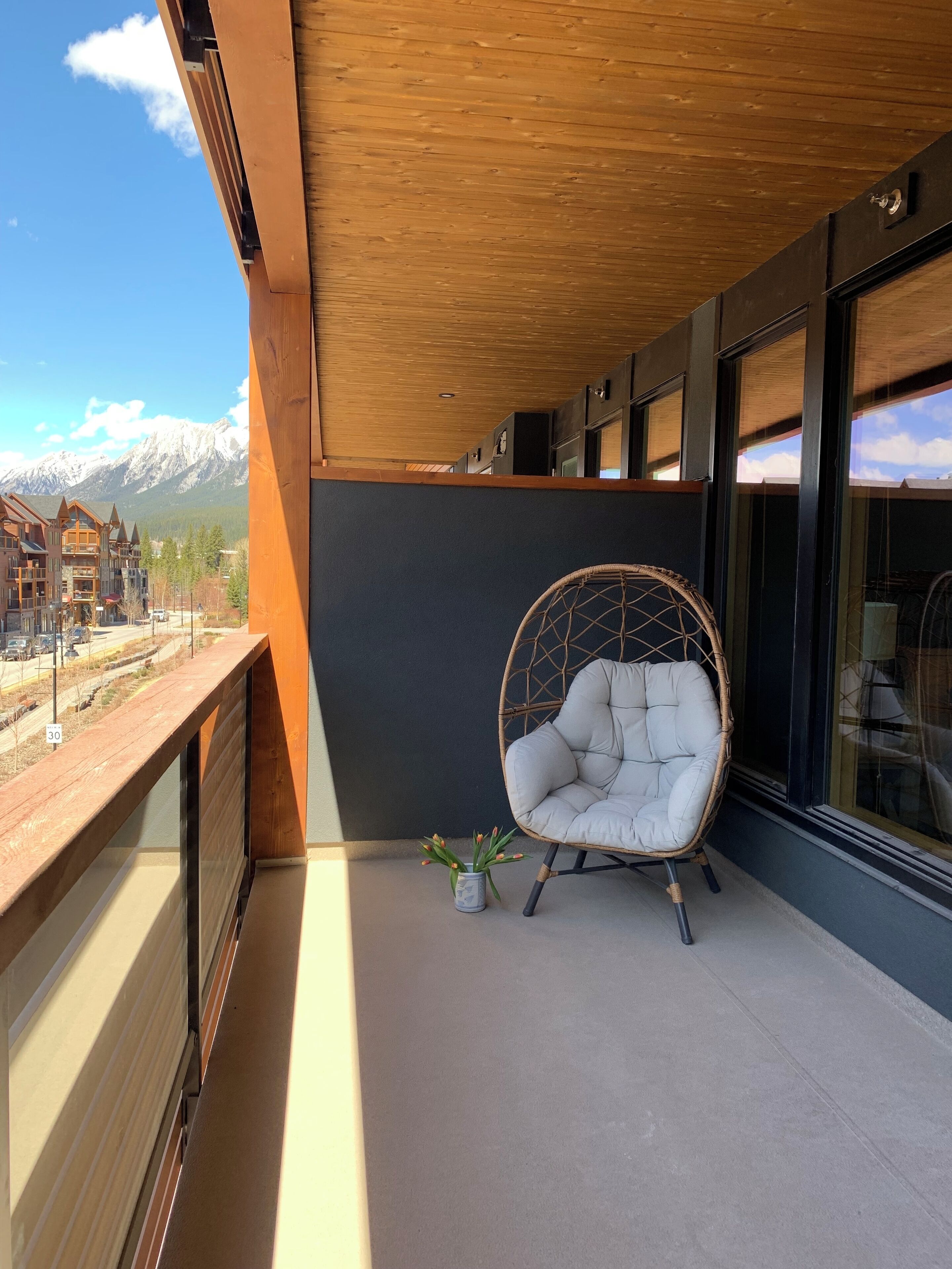A balcony with a wicker chair and a small potted plant, overlooking a mountain range and a town.