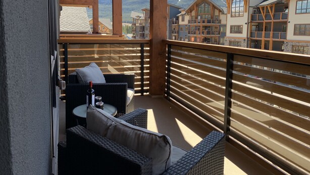 Un balcón con mobiliario de mimbre y una mesa, con vista a un paisaje montañoso y edificios.