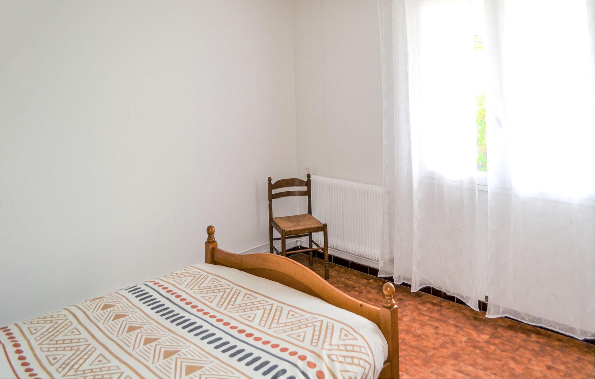 Ein Schlafzimmer mit Bett, Stuhl, Heizkörper und einem Fenster mit durchsichtigen Vorhängen.