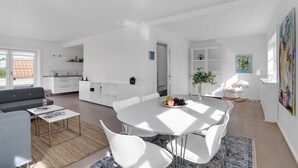 Un espacio moderno y abierto para sala y comedor, con una paleta de colores blanco, una mesa redonda con sillas blancas, un sofá gris y una cocina al fondo.