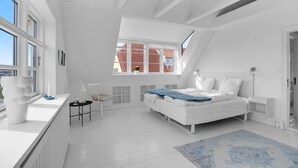 Un dormitorio moderno con decoración en blanco, techo inclinado, cama, escritorio y ventana con vista a un edificio de tejados rojos.