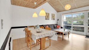 Un coin repas avec une table en bois dressée pour quatre personnes, un canapé avec des coussins et une grande fenêtre donnant sur l’extérieur.