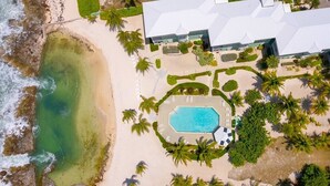 Vista aérea de una propiedad frente a la playa con piscina, palmeras y un edificio grande con techo blanco.