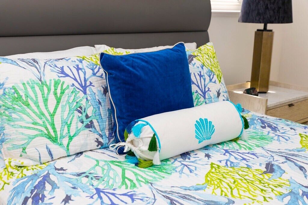 Ein Bett mit floralem Überwurf, einem blauen Kopfkissen und einem gerollten Handtuch mit Muschelmotiv.