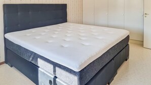 Un lit avec une tête de lit sombre et un matelas blanc, dans une pièce dont le mur est uni et qui possède une porte.