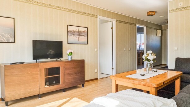 Un salon moderne avec un meuble TV en bois, un téléviseur à écran plat, une table à manger agrémentée d’un vase de fleurs et un canapé gris.