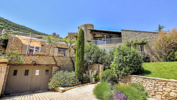 A stone building with a balcony, surrounded by lush greenery and a paved pathway. - Nice home in Condorcet (Condorcet)