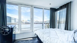 Quarto de hotel moderno com uma cama grande, uma escrivaninha com cadeira e vista para a água e edifícios através de grandes janelas.