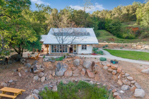 A rustic cabin with a metal roof, surrounded by a natural landscape with trees and a gravel path. - Beautiful, Modern Straw Bale House (Sonora)