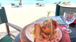 A beachside dining table with a plate of food, a glass of beer, and beach chairs in the background.