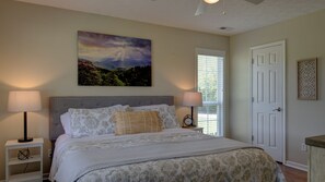 A bedroom with a large bed, two bedside lamps, a clock, a window with blinds, a door, and a landscape painting on the wall.