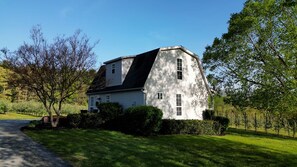 A white two-story house with a steeply pitched roof, surrounded by lush greenery and trees. - Cozy Cottage located in an Apple Orchard - 5 mins from Downtown Hendersonville (Hendersonville)