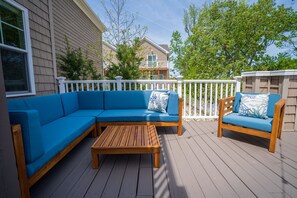 A covered outdoor deck with a blue sectional sofa, a wooden coffee table, and a wooden chair with a blue cushion.