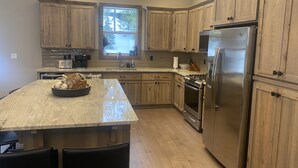 A modern kitchen with wooden cabinets, stainless steel appliances, a large island with seating, and a window providing natural light. - Custom Built Home in 2018 Center of Government Camp (Government Camp)