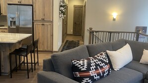 A modern apartment with a grey sofa, a dining area with a refrigerator, and a staircase leading to an upper level. - Custom Built Home in 2018 Center of Government Camp (Government Camp)